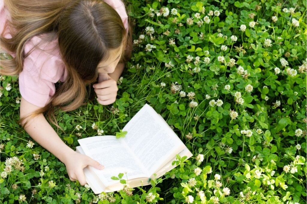 A person with long brown hair lies on green grass and reads an open book, surrounded by small white flowers—a perfect scene to enjoy stories from Free Summer Reading Programs.