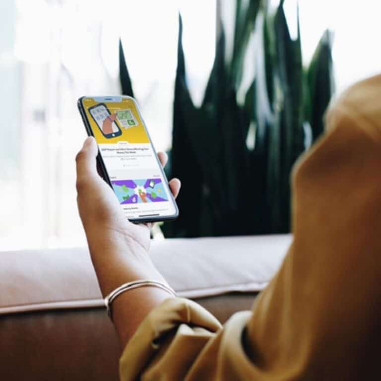 A person is holding a smartphone displaying colorful images and text, showcasing the Easy Investment App offering a $20 Acorns Bonus. The background features a couch and tall green plants.