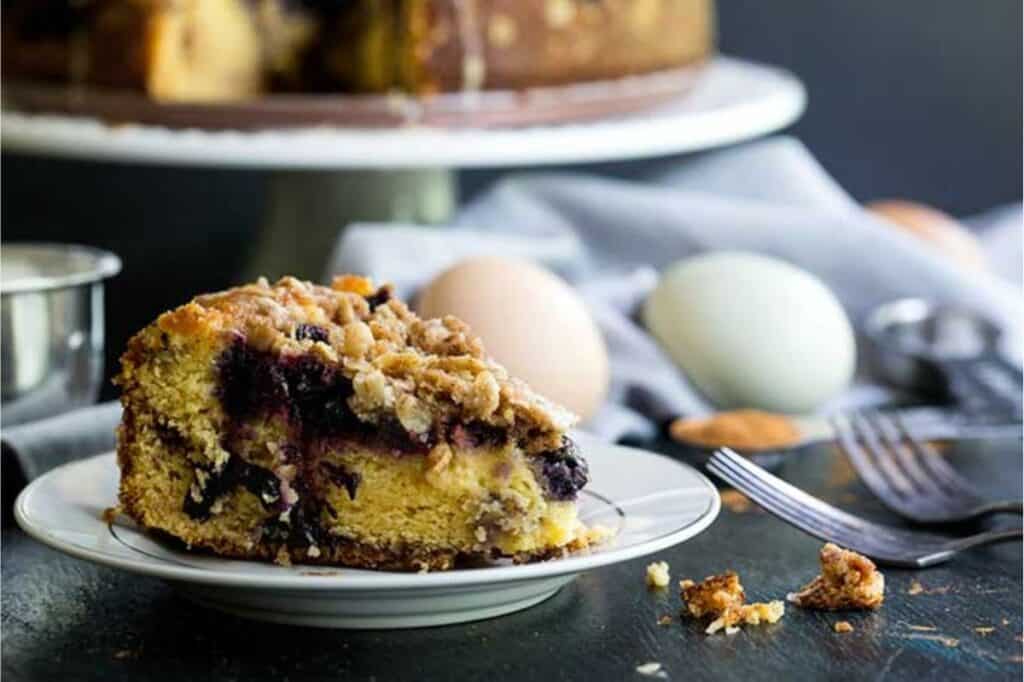 A mouth-watering slice of blueberry crumb cake sits on a white plate with a fork beside it; whole eggs and a cake stand with the remaining cake visible in the background. Try this sweet delight as part of your favorite blueberry recipes.