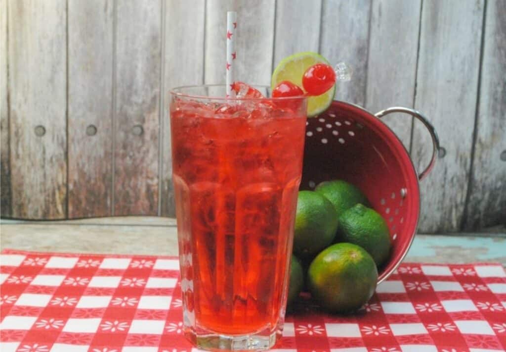 A tall glass of refreshing red iced drink with a straw, garnished with lime and cherries, sits on a checkered tablecloth next to a red colander filled with limes, against a wooden background. Perfect for anyone seeking alcohol-free summer drink recipes to beat the heat!