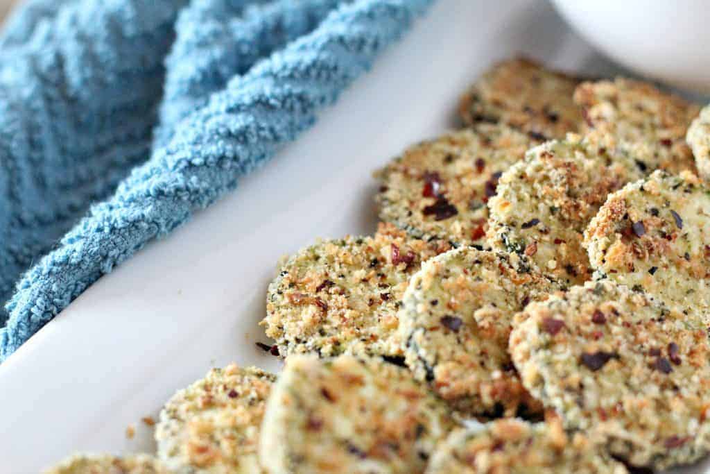 Close-up of a plate with breaded and baked zucchini chips next to a blue textured cloth, showcasing one of the most delicious zucchini recipes.