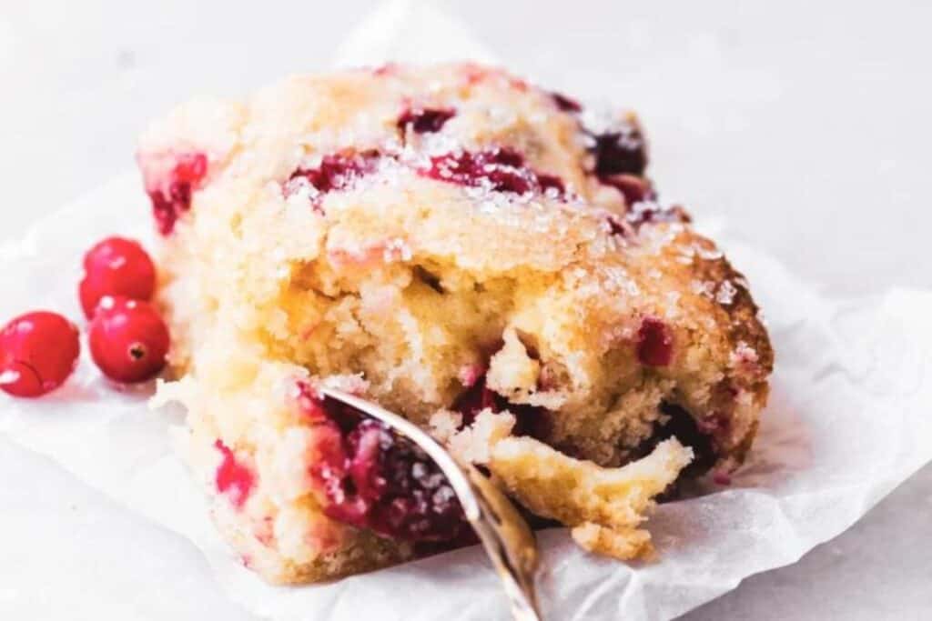 A slice of cranberry cake on a piece of parchment paper with a spoon in it, surrounded by fresh cranberries, ideal for stress-free mornings. Perfect for those seeking easy breakfast recipes.