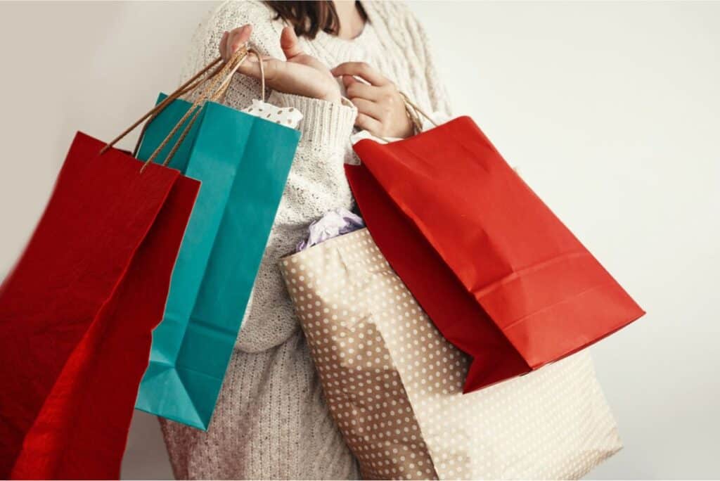 A person in a sweater holds several colorful shopping bags, including red, blue, and polka-dotted ones, against a plain white background, likely planning ahead for Christmas Clearance Sales.