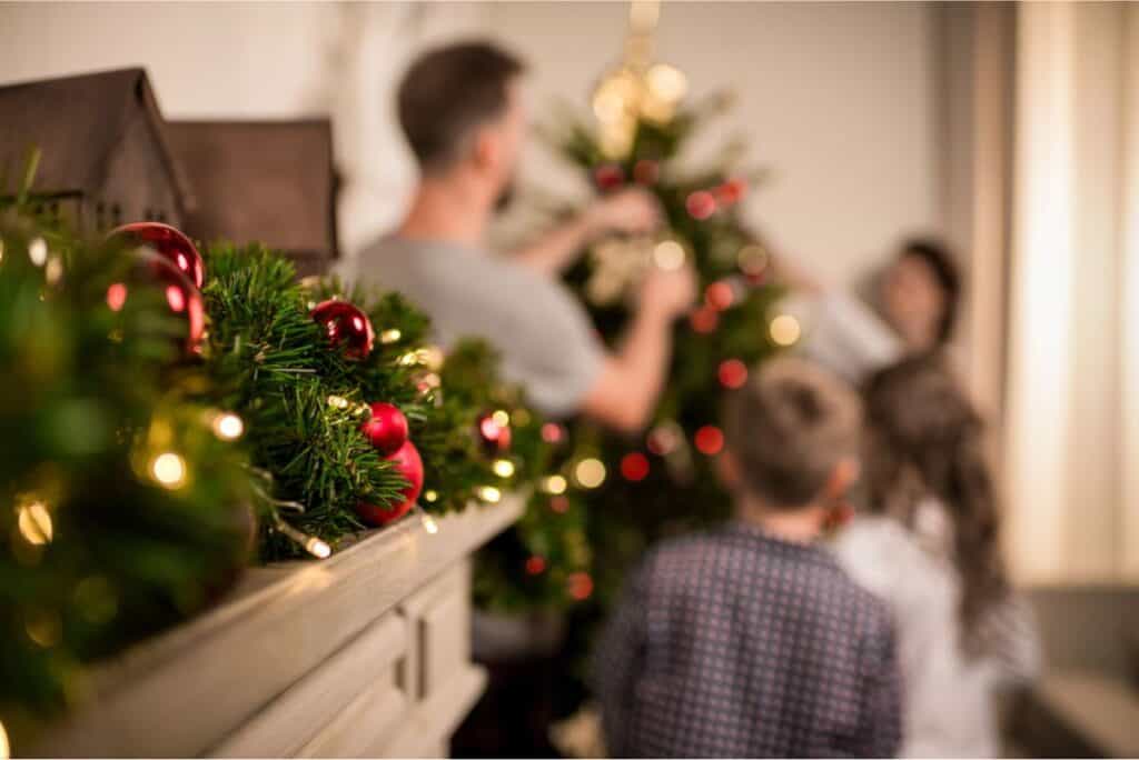 The family is enjoying a stress-free Christmas while decorating a tree in their cozy living room, with a garland adorned with red baubles and lights in the foreground.