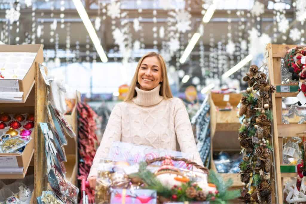 A person wearing a light-colored sweater smiles while standing in a festive aisle filled with holiday decorations, excitedly Planning Ahead for Christmas Clearance Sales.