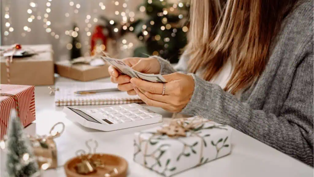 A frugal person counting money at a desk with a calculator, notepad, and wrapped gifts, surrounded by festive holiday decorations.