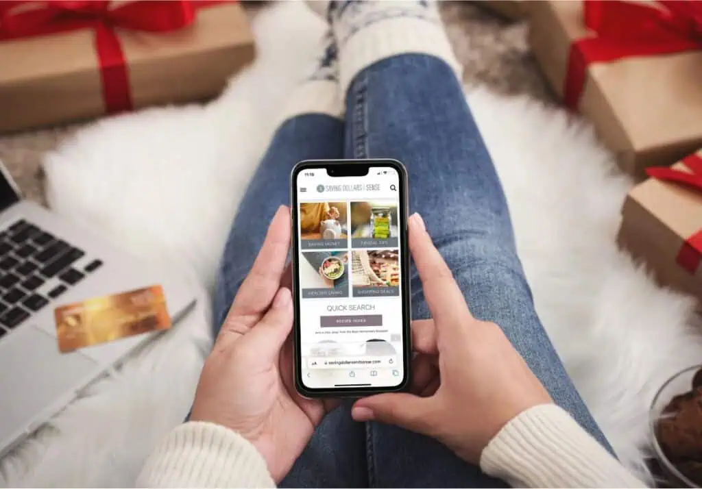 A person in jeans and a white sweater holds a smartphone displaying a recipe website. A laptop, credit card, and gift-wrapped presents are visible in the background, hinting at stay frugal strategies for the holiday season.