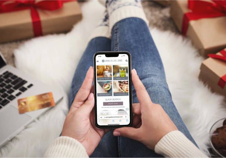 A person in jeans and a white sweater holds a smartphone displaying a recipe website. A laptop, credit card, and gift-wrapped presents are visible in the background, hinting at stay frugal strategies for the holiday season.