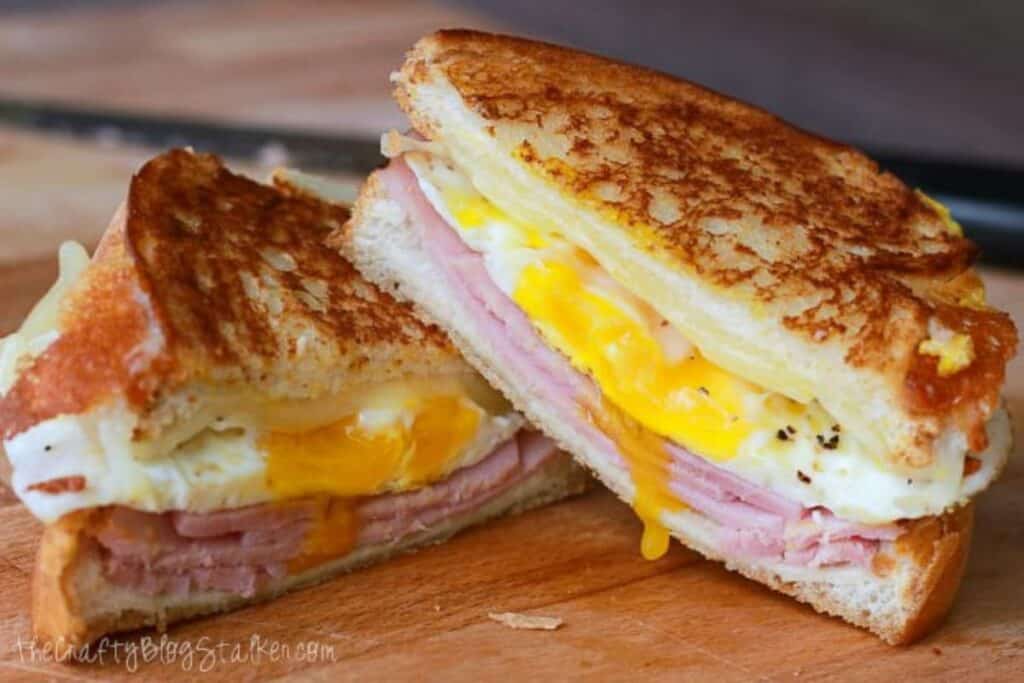 A grilled breakfast sandwich with egg, ham, and melted cheese, sliced in half and placed on a wooden cutting board—perfect for stress-free mornings. For more easy breakfast recipes like this, you'll start every day right!