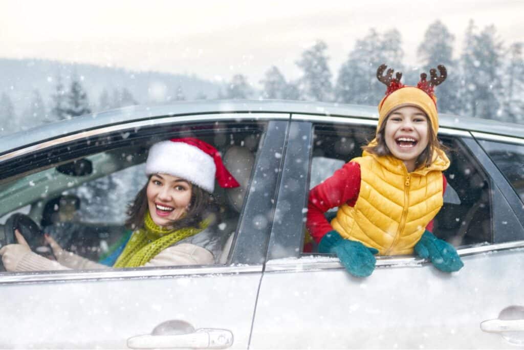 A woman in a Santa hat and a child in reindeer antlers smile out of a car window, embracing the joy of Holiday traveling in a snowy landscape.