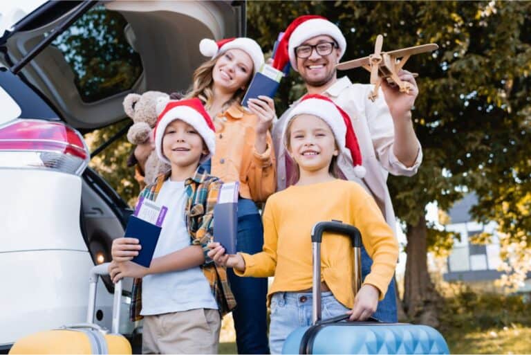 A family of four with Santa hats poses beside a car with an open trunk, holding passports and luggage, indicating preparation for holiday traveling. One man holds a toy airplane, showcasing their excitement while they save money on this festive adventure.