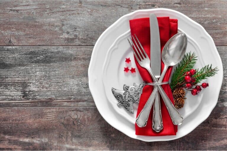 A white plate with a red napkin folded under a fork, knife, and spoon, adorned with small decorative sprigs, berries, and star-shaped accents on a rustic wooden table—perfect for holiday meals.
