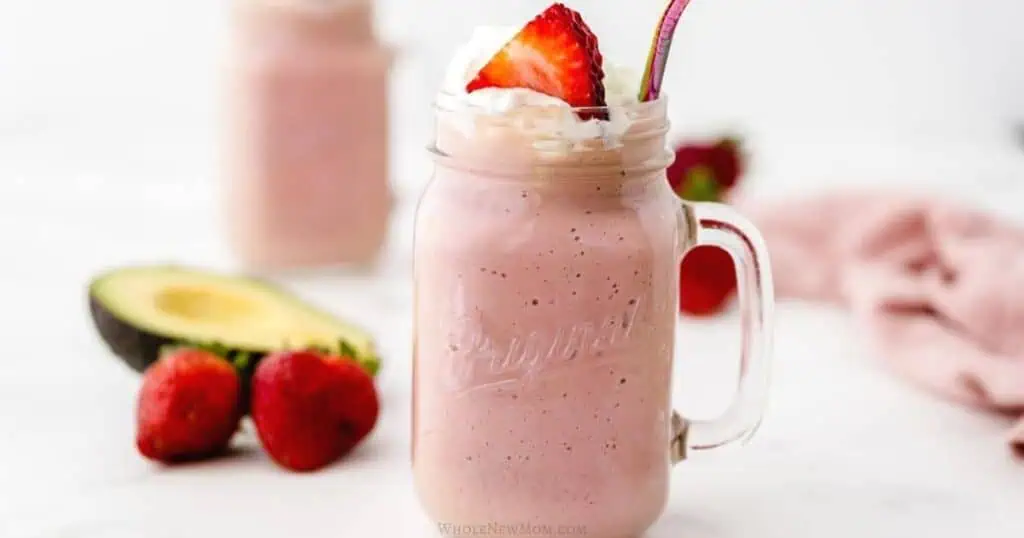 A mason jar filled with a pink smoothie topped with whipped cream and a strawberry slice, with a metallic straw. Nearby, a halved avocado and whole strawberries are placed on a white surface, offering perfect breakfast ideas for stress-free mornings.