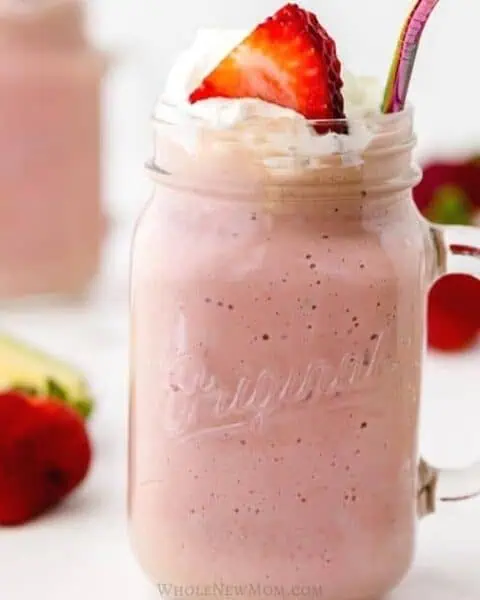 A mason jar filled with a pink smoothie topped with whipped cream and a strawberry slice, with a metallic straw. Nearby, a halved avocado and whole strawberries are placed on a white surface, offering perfect breakfast ideas for stress-free mornings.