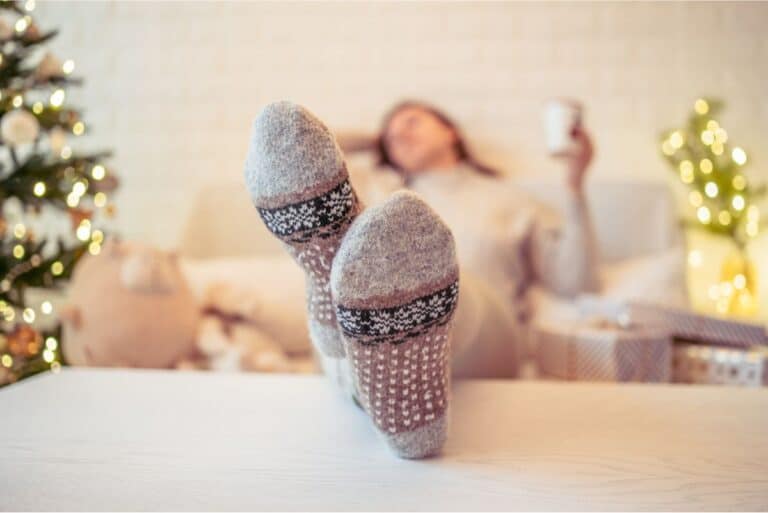 Person wearing grey patterned socks, relaxing on a couch with a Christmas tree in the background, enjoying holiday stress management with a mug in one hand.