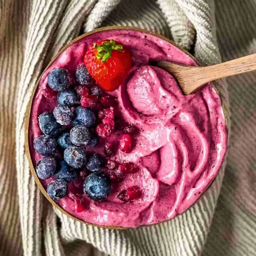 A bowl of pink smoothie topped with blueberries, raspberries, and a whole strawberry, placed on a textured fabric. A wooden spoon rests in the smoothie. Perfect for stress-free mornings, this delightful dish is a champion of easy breakfast recipes.