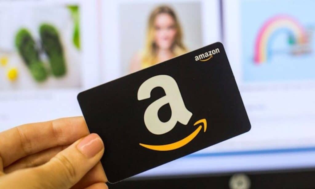 A hand holds an Amazon gift card with the iconic white "a" logo and orange arrow, ready for a giveaway, against a blurred background of images on a screen.