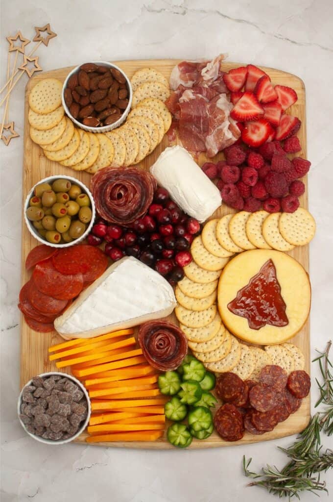 A festive charcuterie board featuring an array of cheeses, meats, crackers, olives, and fresh strawberries and raspberries adds a touch of Christmas magic. Delight in the addition of chocolate chips and almonds, all beautifully arranged on a rustic wooden board.