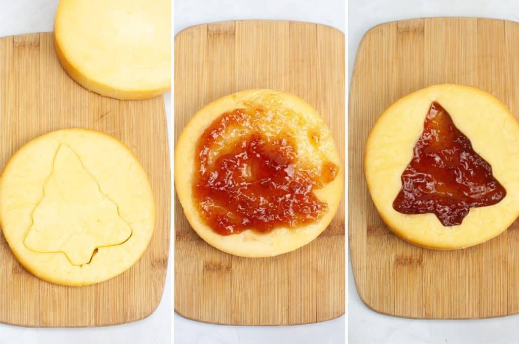 A round cheese wheel rests on a wooden board, featuring a tree-shaped cutout. Jam is spread on one side while filling the cutout on the other, creating a festive Christmas charcuterie centerpiece.