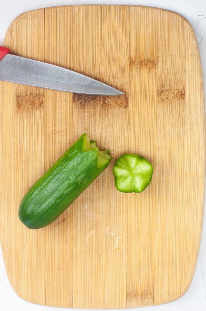 A partially sliced cucumber rests on a wooden charcuterie board, with a knife placed beside it, ready to add a touch of freshness to your Christmas spread.