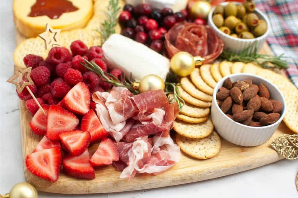 A festive charcuterie board perfect for Christmas, featuring strawberries, raspberries, prosciutto, almonds, crackers, olives, cured meat, cheese, cranberries, and rosemary sprigs.