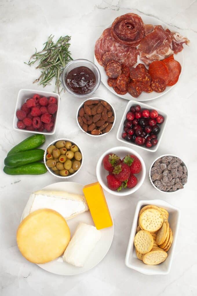 A festive charcuterie board featuring cured meats, cheeses, crackers, olives, almonds, and berries is complemented by chocolate chips and cranberries. Topped with a rosemary garnish, it's the perfect centerpiece for your Christmas gathering.