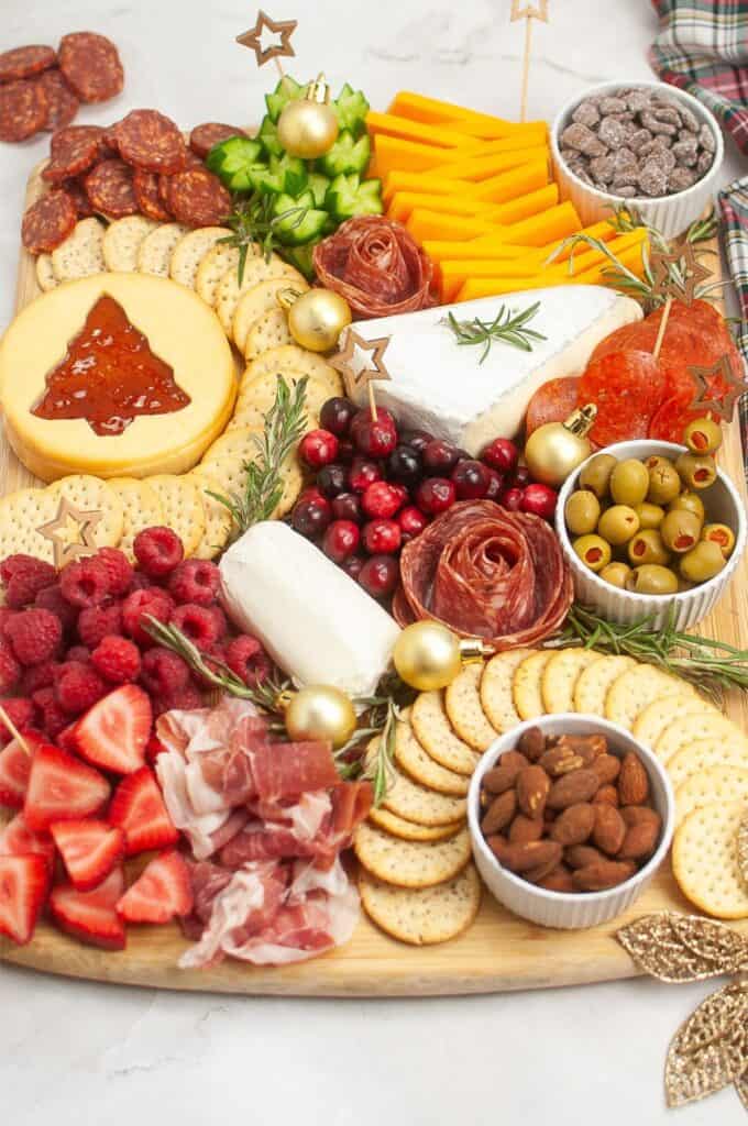 A festive charcuterie board beautifully arranged with cheeses, crackers, olives, nuts, raspberries, and more is adorned with Christmas decorations and rosemary sprigs.