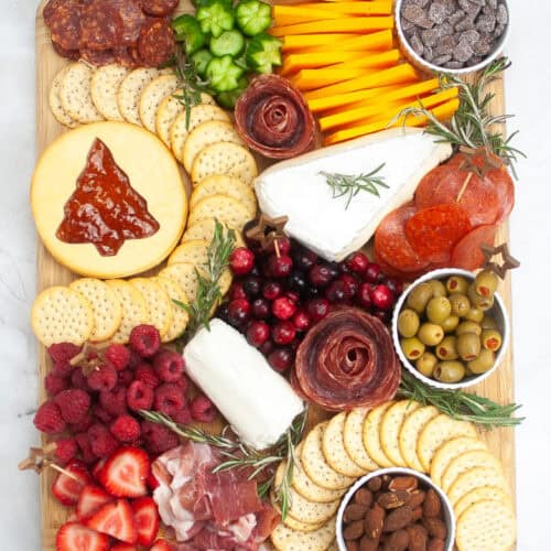 A festive Christmas charcuterie board featuring cheeses, crackers, meats, and an array of raspberries, strawberries, cranberries. Adorned with star-shaped cucumbers, chocolate pieces, olives, almonds and garnished with sprigs of rosemary.