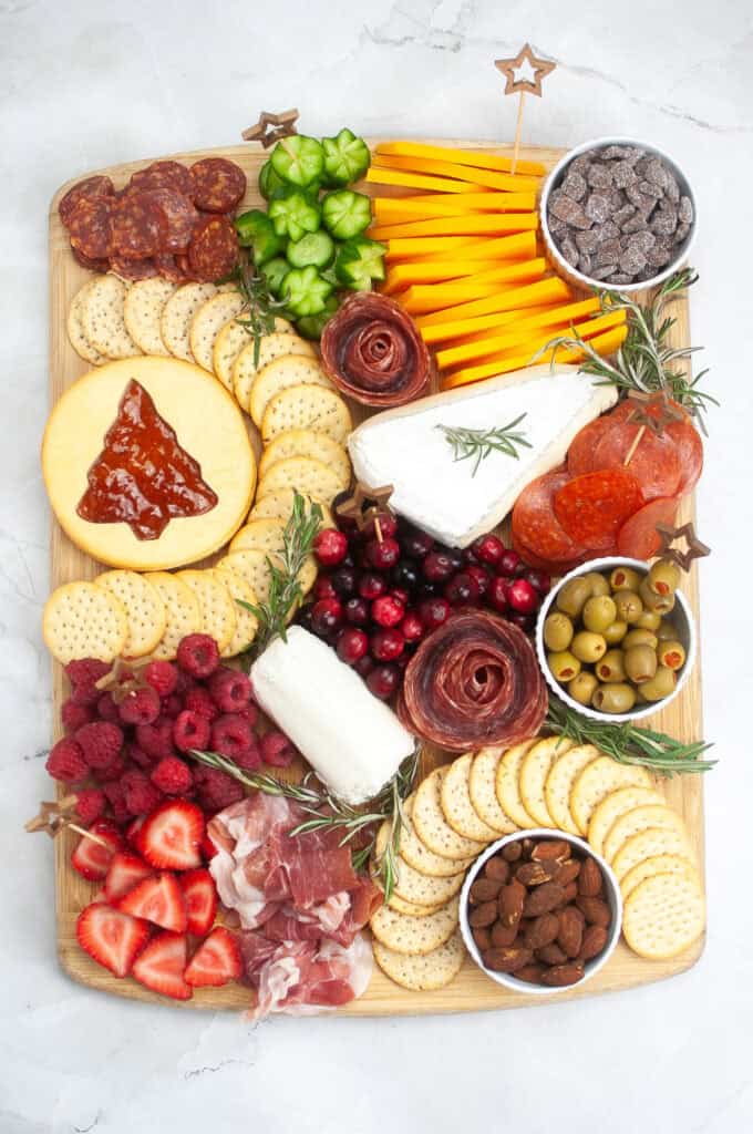A festive Christmas charcuterie board featuring cheeses, crackers, meats, and an array of raspberries, strawberries, cranberries. Adorned with star-shaped cucumbers, chocolate pieces, olives, almonds and garnished with sprigs of rosemary.