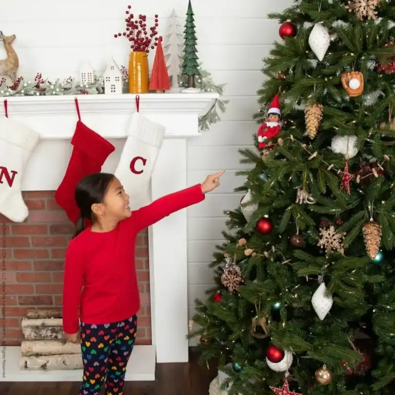 A young girl in festive attire excitedly points at a decorated Christmas tree next to a fireplace with stockings, while an Elf on a Shelf playfully peeks from its spot.
