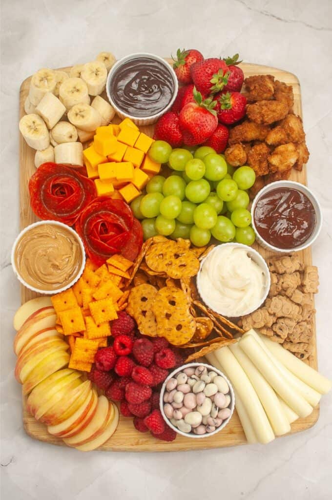 A delightful kids charcuterie board adorned with a wooden platter brimming with bananas, strawberries, grapes, apples, raspberries, cheese cubes, pretzels, crackers, breaded chicken, dip bowls, and candies.