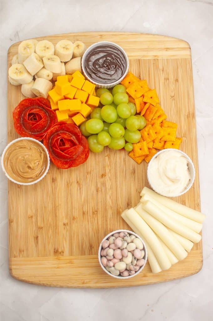 A delightful kids charcuterie board features a wooden platter with neatly arranged banana slices, cheese cubes, charming pepperoni roses, juicy grapes, crunchy crackers, kid-friendly dipping sauces, fun string cheese shapes, and colorful coated candies.