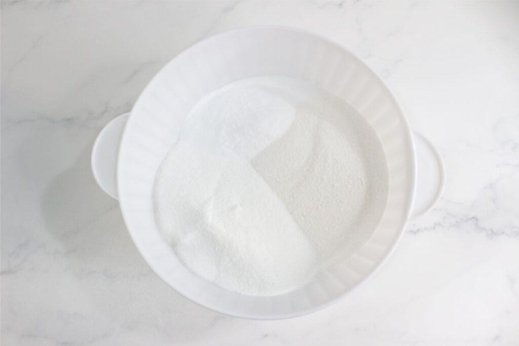 A white mixing bowl filled with fine homemade granulated sugar sits on a white marble surface.