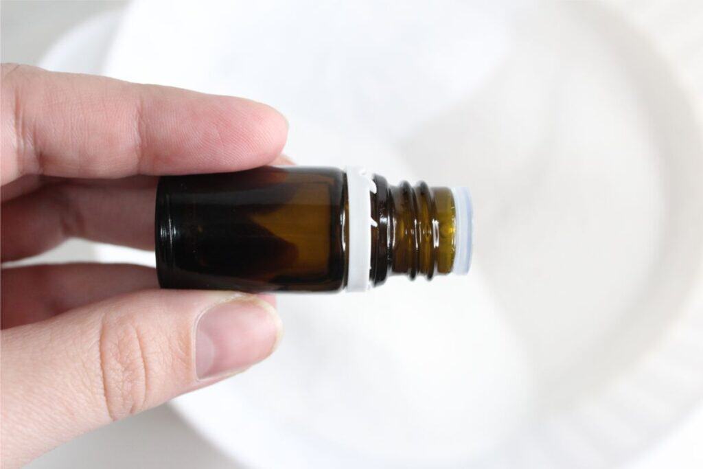 A hand holds a small, dark amber glass bottle with an open top, positioned over a white surface. The bottle, perfect for storing homemade remedies or natural laundry solutions, captures an essence of simple elegance and functionality.