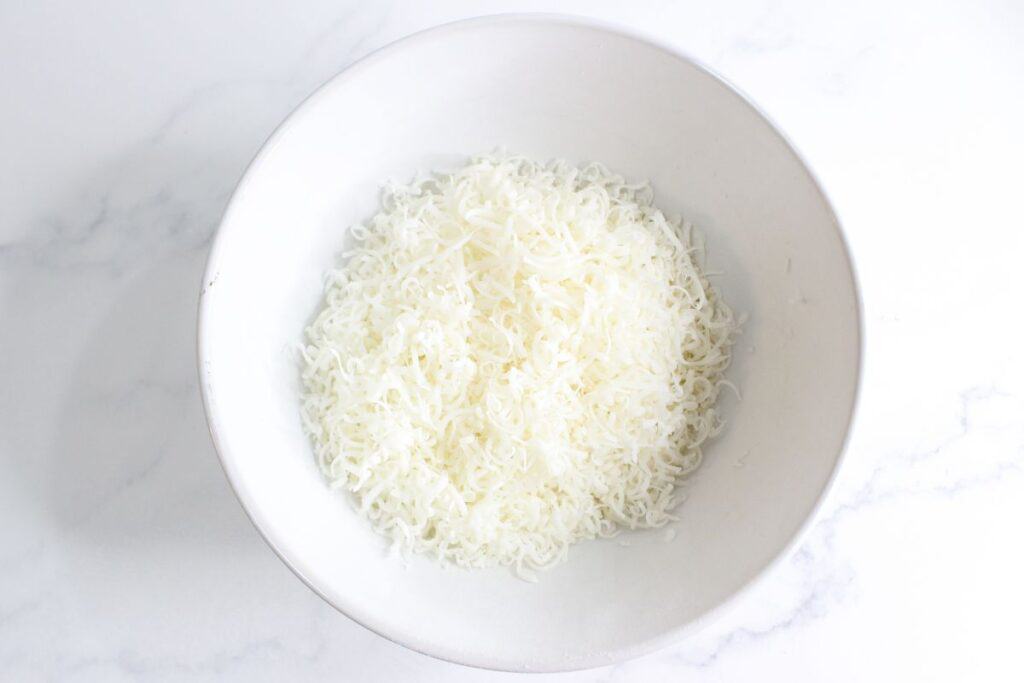 A bowl filled with homemade shredded mozzarella cheese on a white marble surface.