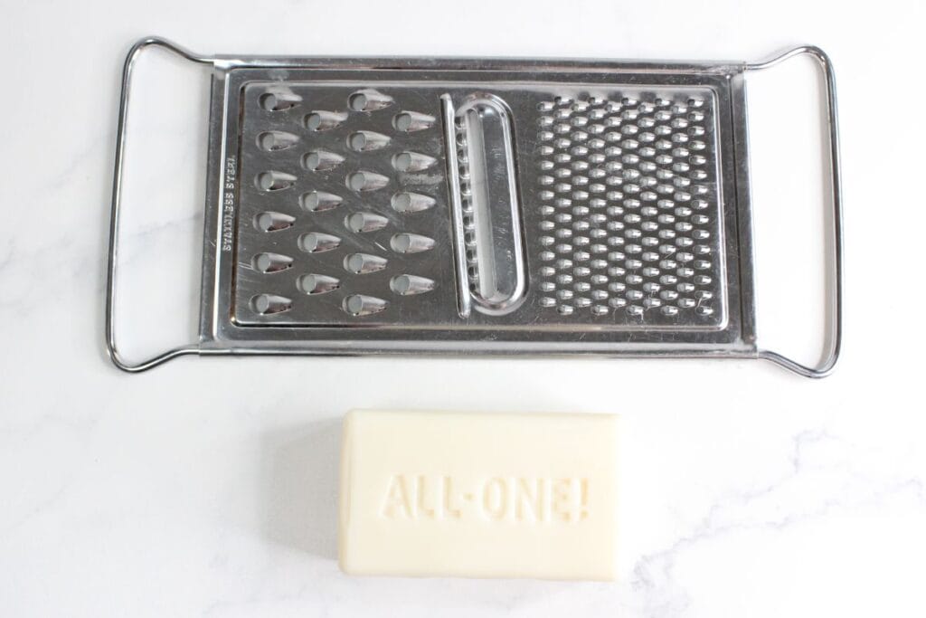 A metal cheese grater and a bar of soap with "ALL-ONE!" embossed on it are placed on a white marble surface, hinting at the creation of homemade laundry powder.