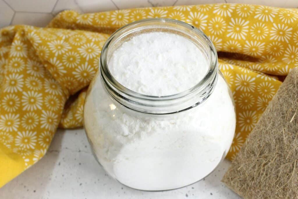 A glass jar filled with homemade white powder, possibly baking soda or flour, sits on a white speckled surface with a yellow floral cloth in the background, evoking the simplicity of crafting your own laundry powder.