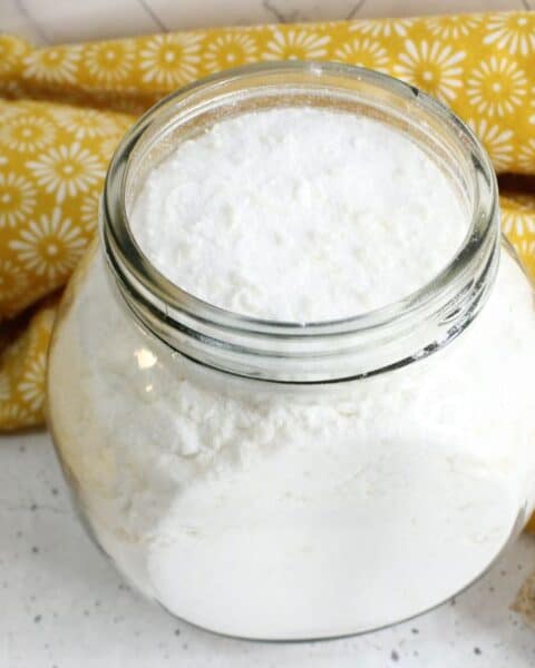 A glass jar filled with homemade white powder, possibly baking soda or flour, sits on a white speckled surface with a yellow floral cloth in the background, evoking the simplicity of crafting your own laundry powder.