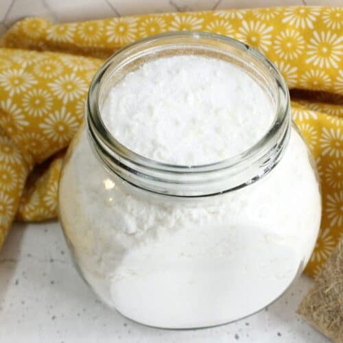 A glass jar filled with homemade white powder, possibly baking soda or flour, sits on a white speckled surface with a yellow floral cloth in the background, evoking the simplicity of crafting your own laundry powder.