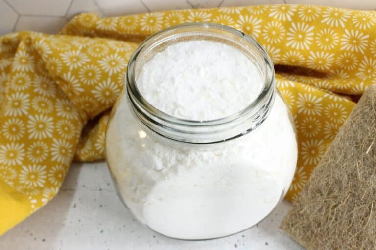 A glass jar filled with homemade white powder, possibly baking soda or flour, sits on a white speckled surface with a yellow floral cloth in the background, evoking the simplicity of crafting your own laundry powder.