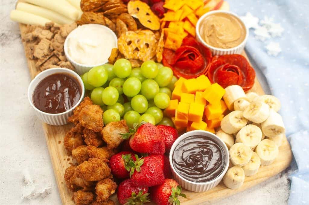 A delightful kids charcuterie board featuring a wooden platter filled with an assortment of snacks like grapes, strawberries, bananas, cheese cubes, pretzels, cookies, chicken bites, and various dipping sauces. Perfect for little ones' appetites and adventurous taste buds!.