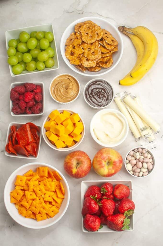 Create a delightful kids charcuterie board with an array of snacks, featuring juicy grapes, crunchy pretzels, bananas, and sweet raspberries. Add a touch of peanut butter and chocolate spread alongside cheese, hummus, yogurt-covered raisins, savory crackers, apples, strawberries, pepperoni slices, and string cheese.