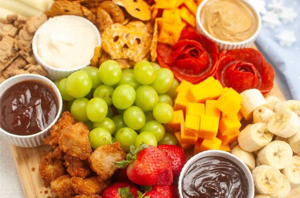 A delightful kids charcuterie board featuring a snack platter with grapes, strawberries, bananas, cheese cubes, pretzels, pepperoni roses, chicken bites, and assorted dips.