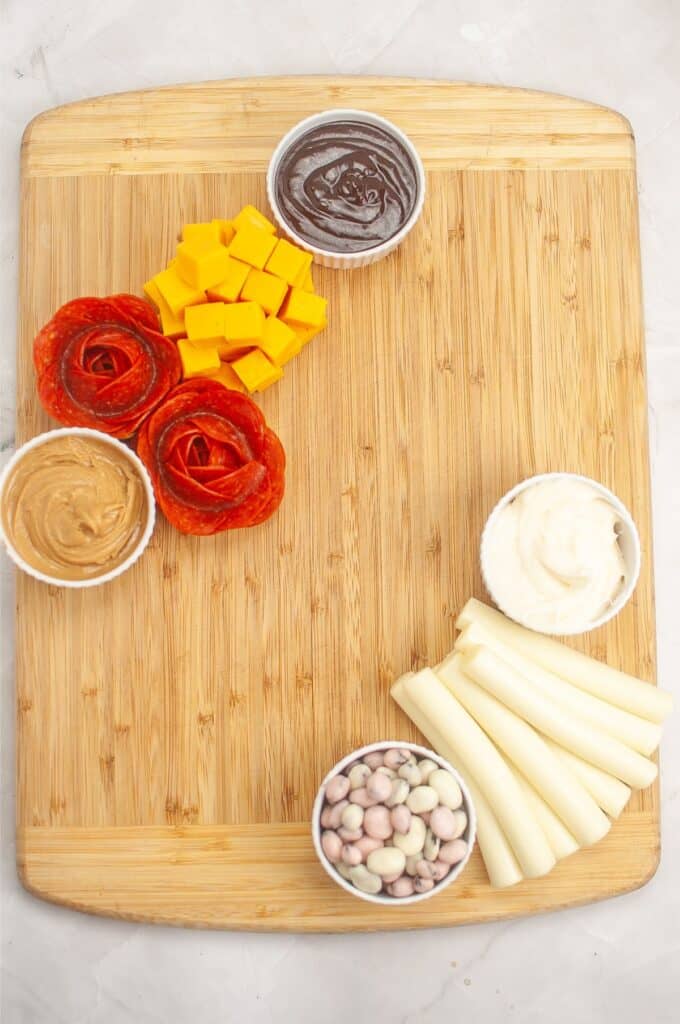 A delightful kids' charcuterie board featuring a wooden board with pepperoni slices, cheese cubes, chocolate spread, peanut butter, white dip, string cheese, and yogurt-covered treats in small containers.