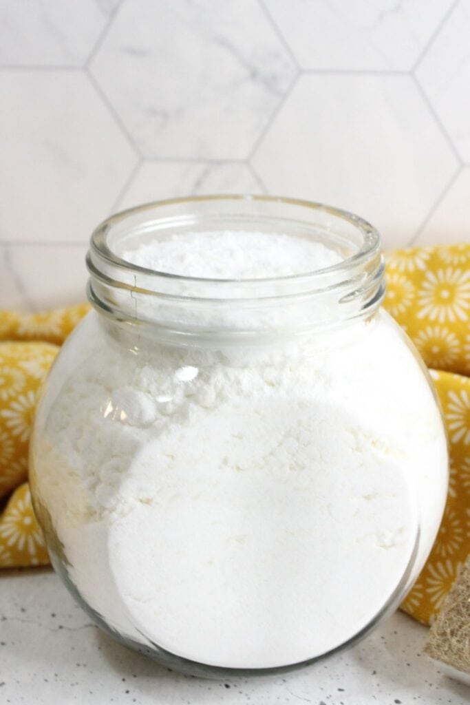 A glass jar filled with homemade laundry powder sits on a speckled counter, complemented by a yellow patterned cloth in the background.