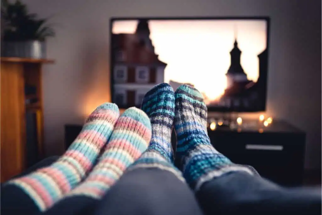 Two people sporting colorful knitted socks relax on a couch, streaming their favorite show on a TV that displays a cityscape at sunset, taking advantage of Cyber Monday deals.