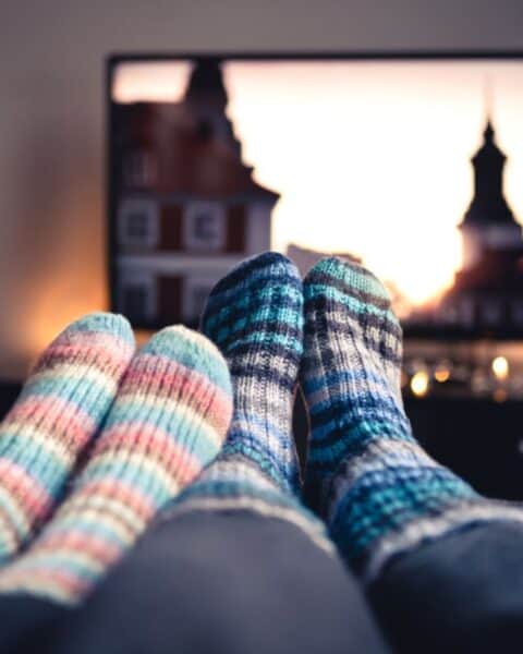 Two people sporting colorful knitted socks relax on a couch, streaming their favorite show on a TV that displays a cityscape at sunset, taking advantage of Cyber Monday deals.