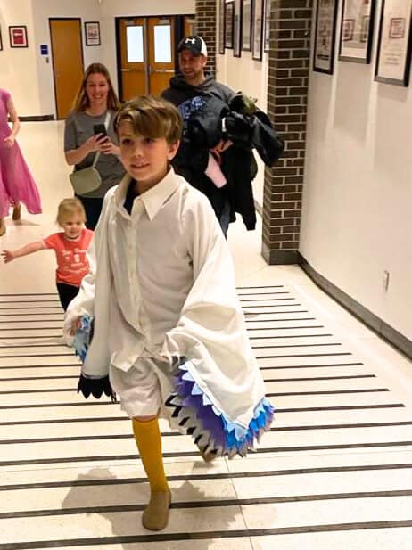 Liam Takes the Stage in a white costume with blue accents, walking down the hallway, followed by smiling adults and a child.