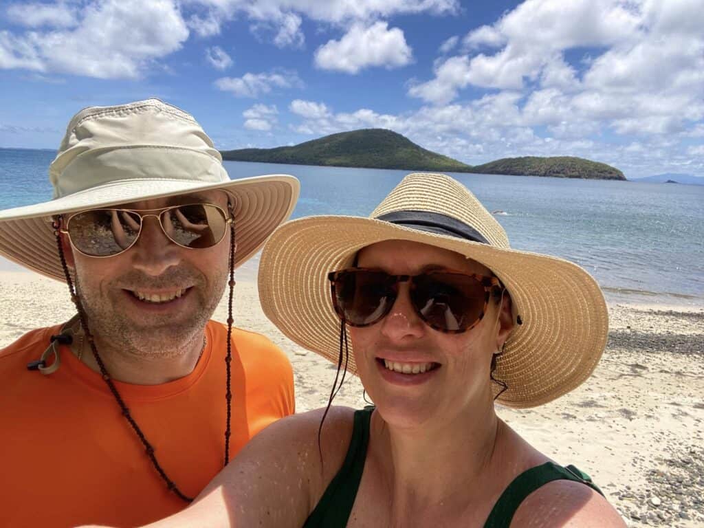 Two people wearing sun hats and sunglasses are smiling under the bright sunshine on a beach. The ocean and small islands are visible in the background under a partly cloudy sky, offering a picturesque escape from boring ordinary life.