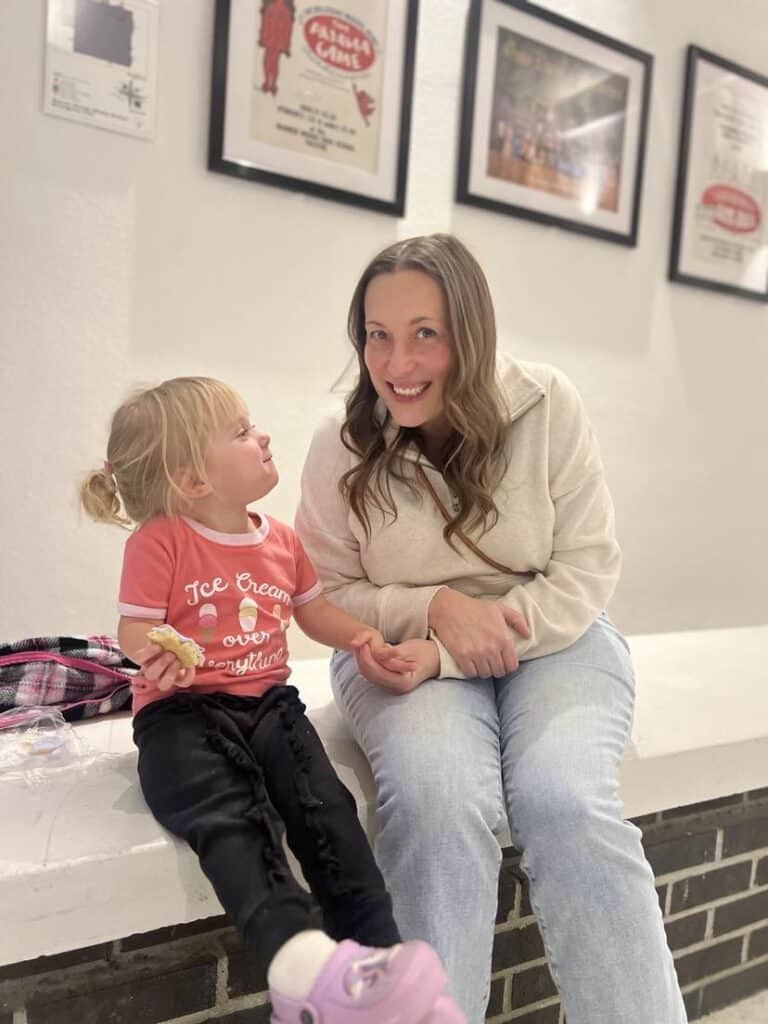 A woman and a child sit on a white bench indoors. The child, wearing a red shirt and holding his favorite snack, smiles as the woman beams at the camera. Framed pictures of their favorite things hang on the wall behind them, creating a cozy backdrop for this cherished moment.